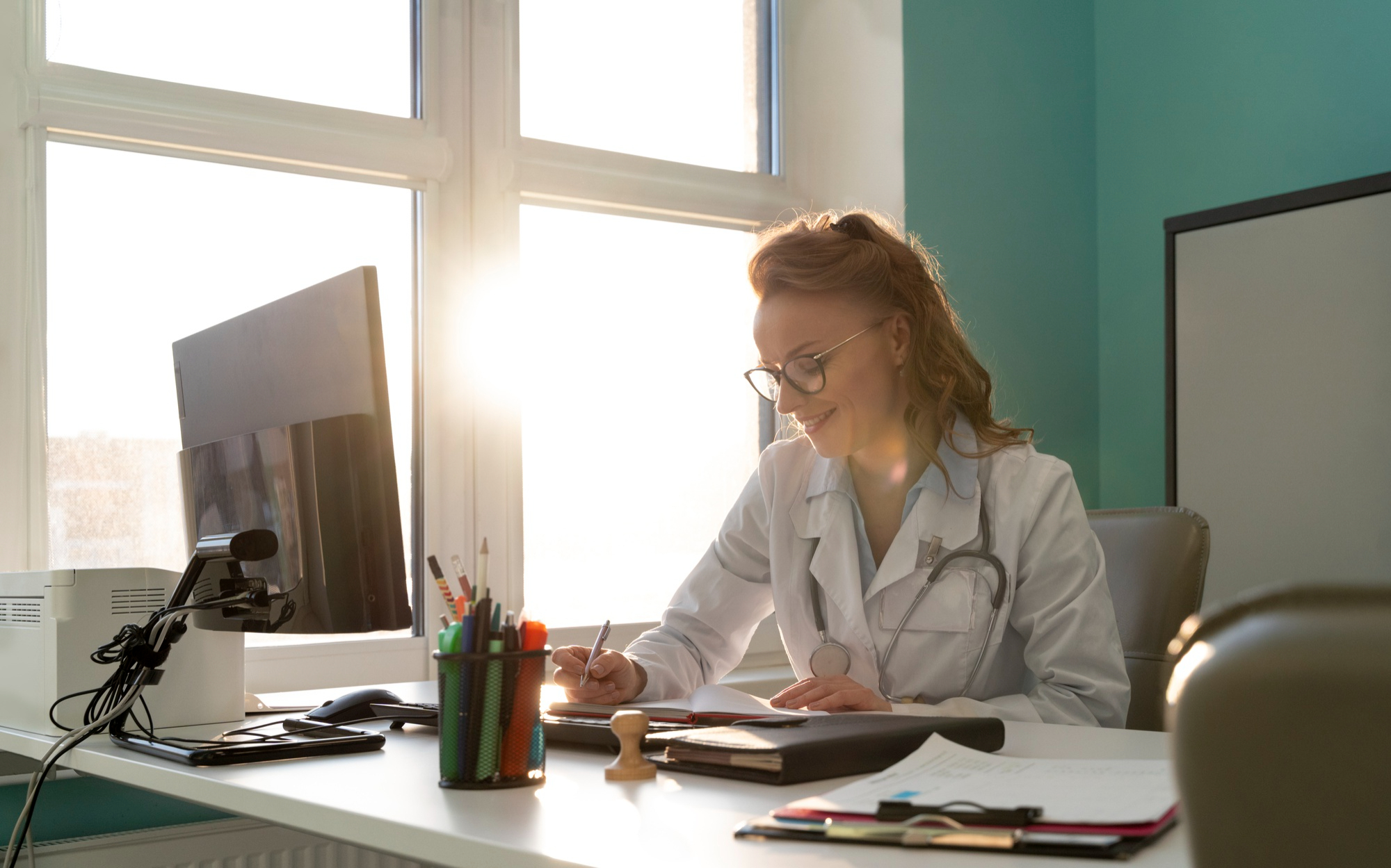 Secretária médica sentada na frente de um computador, ao lado de uma janela, escrevendo dados.